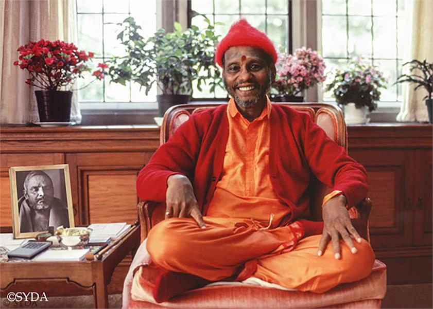 Baba sitting cross legged on a chair, smiling, next to a photo of Nityananda,