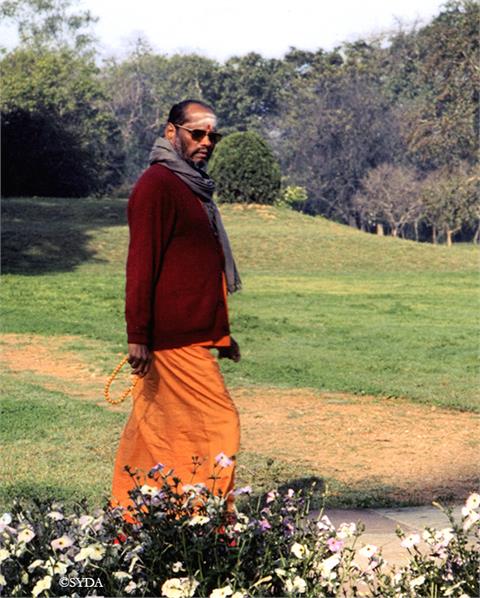 Baba walking through gardens with sunglasses, holding a japa mala