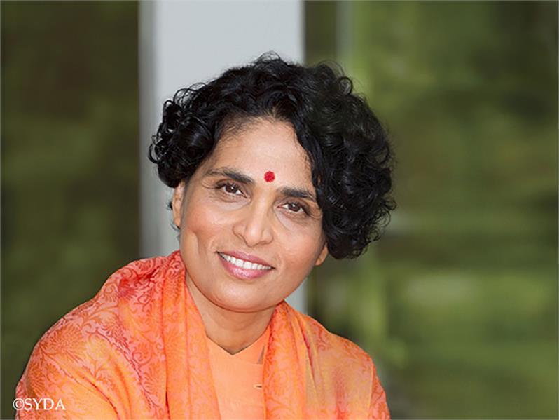 Close up of Gurumayi smiling in front of a window with green foliage in the background