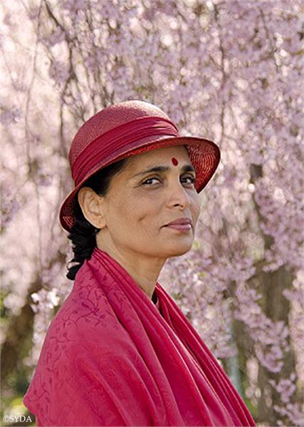 Gurumayi in front of hanging pink flowers with red robes and hat