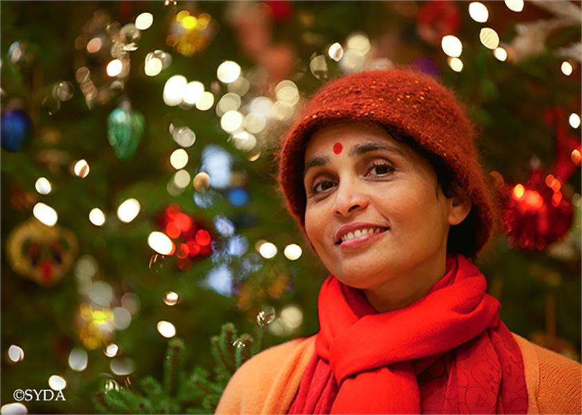 Gurumayi in front of a blurred christmas tree with lights, wearing a fluffy red hat and scarf