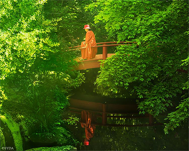 Wide shot of Gurumayi on a bridge over a lake, surrounded by bright green foliage