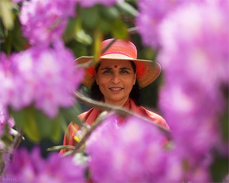 Close up of Gurumayi in a wide brimmed hat,  standing behind a ring of purple flowers