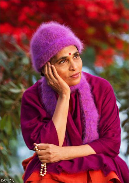 Gurumayi resting her hand on her head in pink and purple clothes, against a nature background