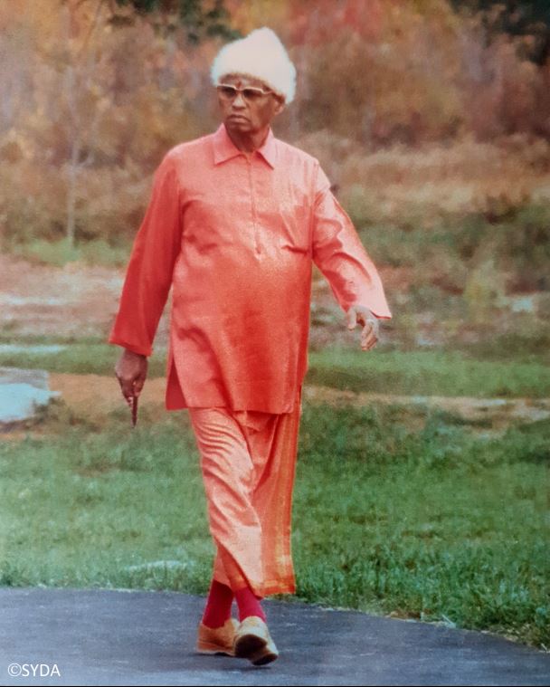 Full body photo of baba walking outside in orange robes, white hat and sunglasses