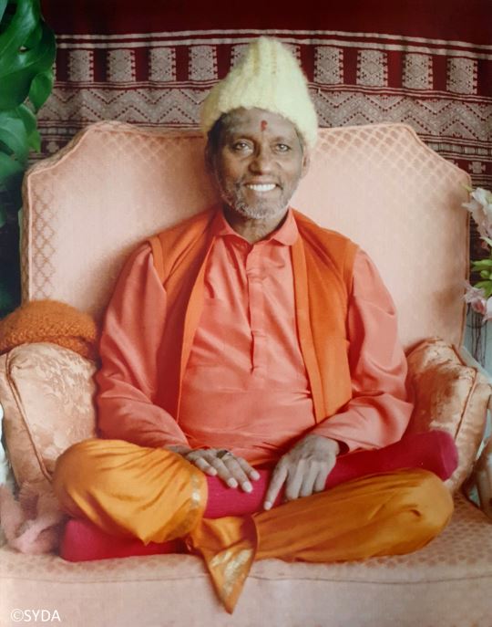Baba sitting cross legged on chair, smiling, with white hat